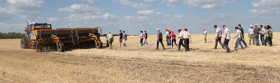 Russia People walking across a harvested field near large farming machinery under a blue sky with scattered clouds